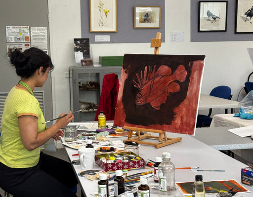 A person in a yellow shirt sits at a white table cluttered with paint tubes, brushes, and art supplies, painting on a canvas with a reddish-brown abstract design in an art studio.
